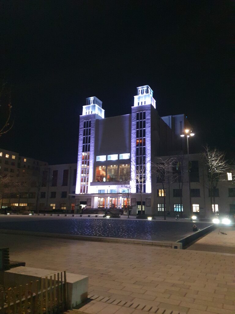 he plaza and theater of Villeurbanne,  there is a square water basin in the middle, no water at the moment, but in summer it is full of playing children. beyond the theater a square white sovietic styles building with a bgger part in the middle with 2 square "towers". It is night, everythinf is mostly in the dark, except the theater, illumuniated in white / purple.