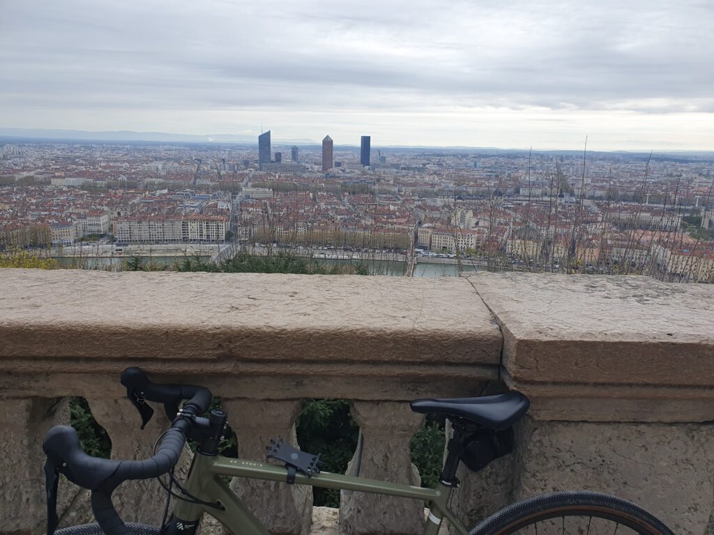 picture of a gravel bike resting on a stone parapet. beyond the rooftops of Lyon, with tower of business center in the middle, the Alps far away, and lots of grey clouds in the sky