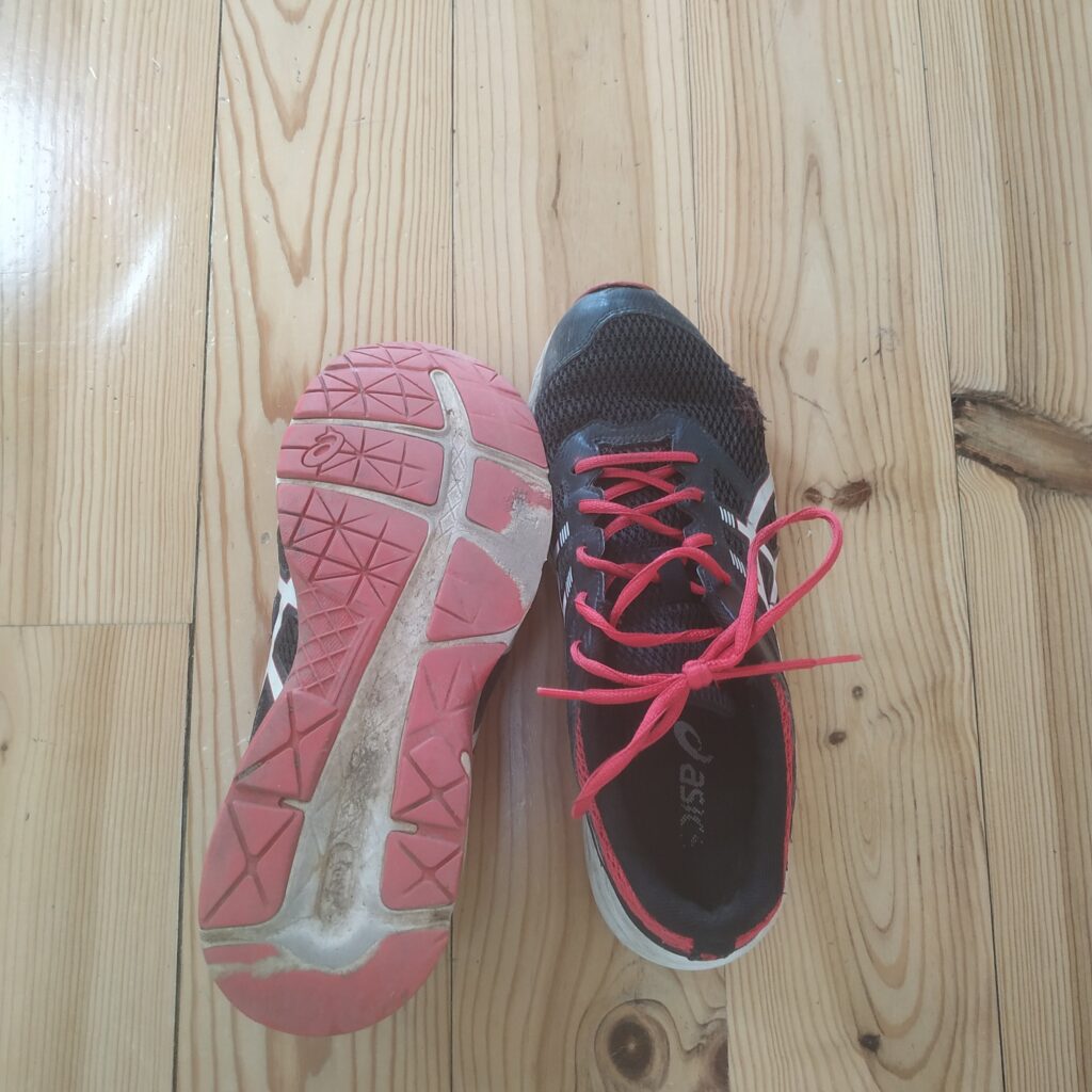 a pair of very tired pair of running shoes, from above. on the left we see the worn out sole, on the right the ripped and repaired mesh. shoes are black, with red sole and laces.