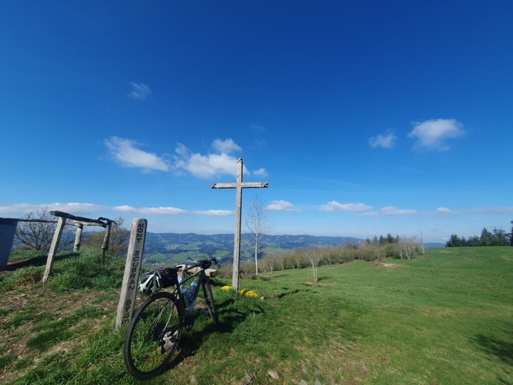 Bike resting on Soubran summit sign, with cross nearby, hills beyond, an ddeep blue sky above