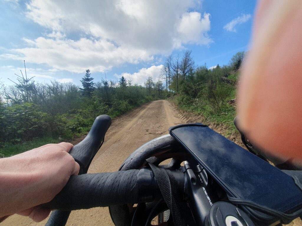 awkward picture taken while riding a hardpack trail. we can see my left hand holding handlebar, the trail going up, bordered by llow bushes, blue sky with a big white cloud above. small part of picture covered by a finger.