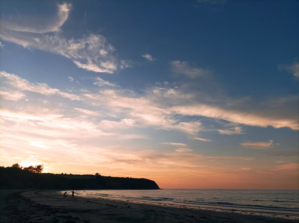 Night view from the beach, of the sea and the dark cliff beyond which the sun is setting, casting an orange / pink glow. Sky ladded with some streamy clouds